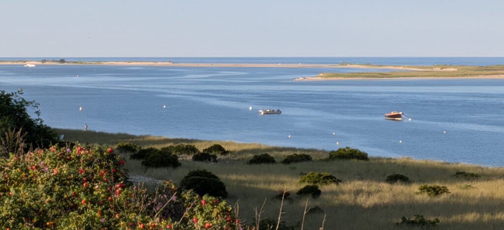 New England coastal harbor with dunes and boats at sunset.