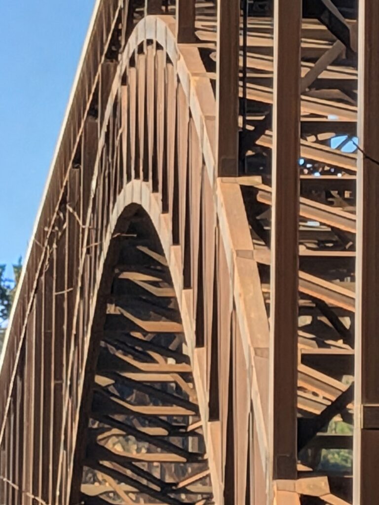 A close-up, upward-angled view of a large, rust-colored steel arch bridge. The intricate lattice of beams and supports is visible against a clear blue sky, creating a pattern of light and shadow.